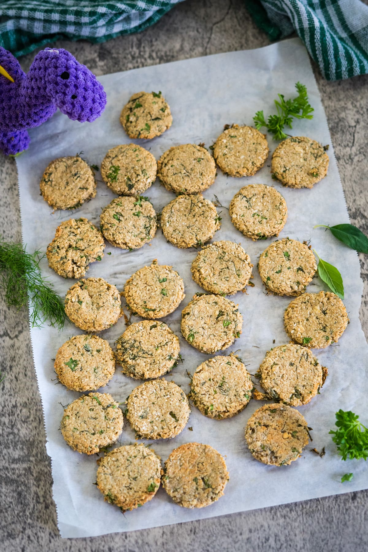 A sheet of freshly baked round cookies with herbs, arranged in rows on parchment paper, with some green garnishes and a purple knit toy on the side.