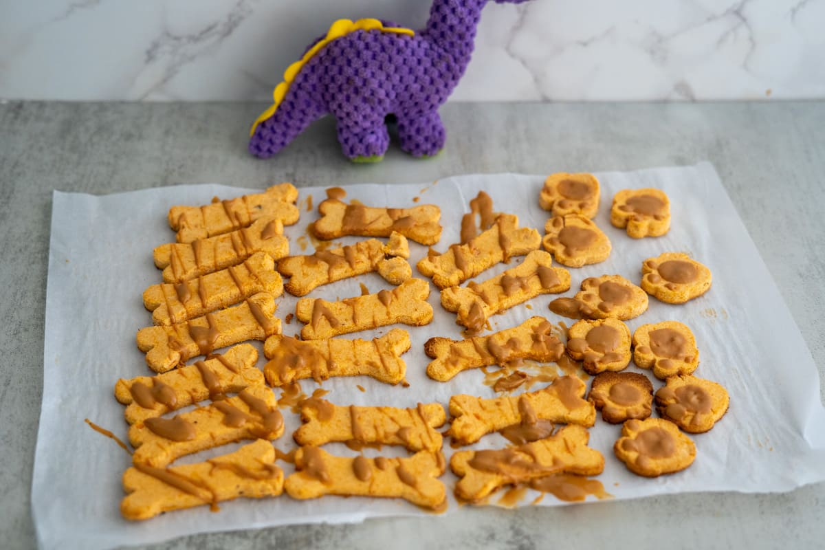 Baked dog treats, shaped like bones and paws, are arranged on parchment paper. A small purple dinosaur toy is in the background.