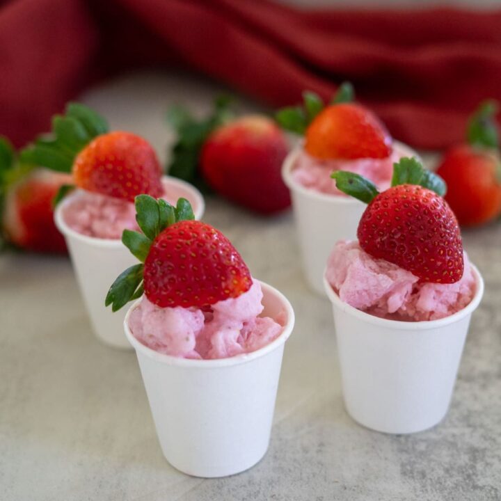 Four small white cups filled with pink strawberry mousse, each topped with a whole strawberry, placed on a light surface. Red fabric and strawberries in the background.