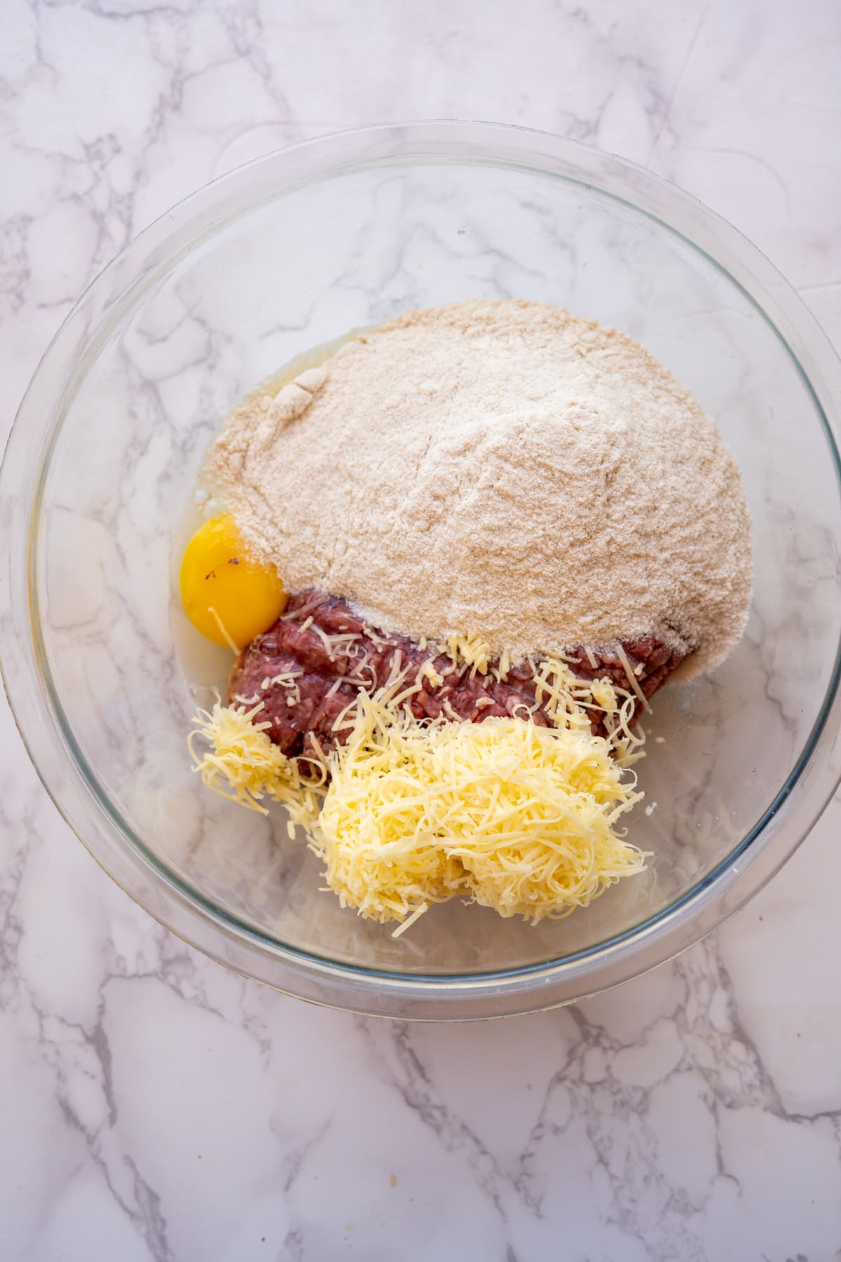 Ingredients in a glass bowl: ground beef treats for dogs, shredded cheese, a cracked egg, and a heap of breadcrumbs on a marble countertop.