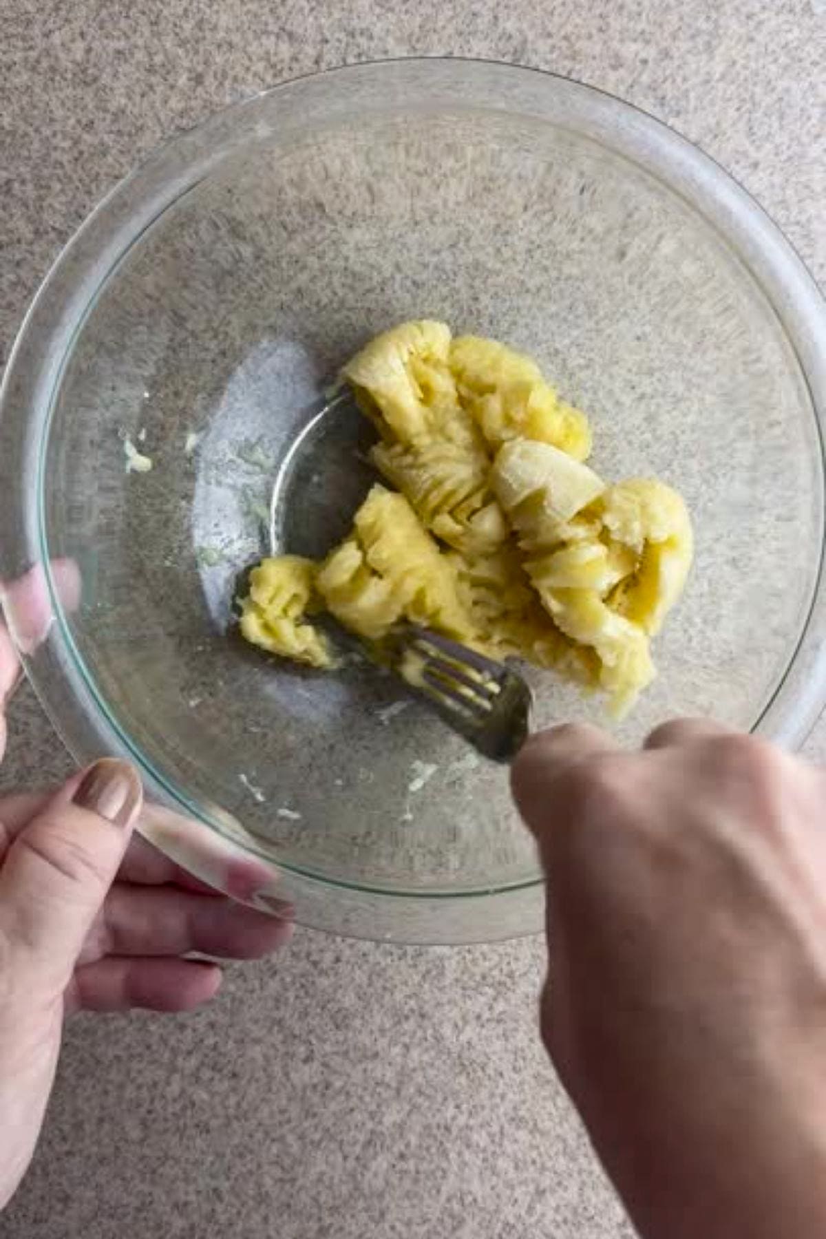 A person mashes bananas with a fork in a clear glass bowl on a kitchen countertop.