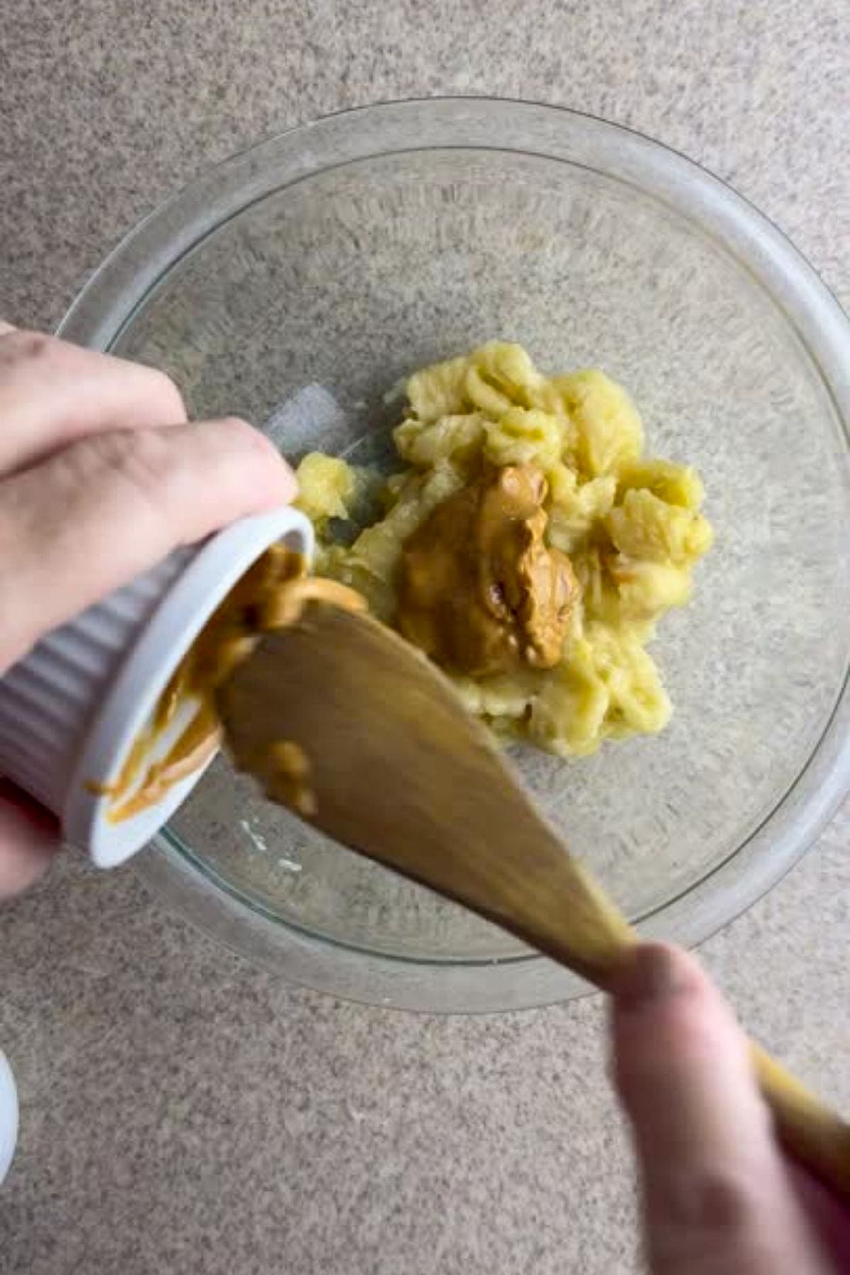 A person is adding peanut butter to a bowl of mashed bananas using a wooden spatula.