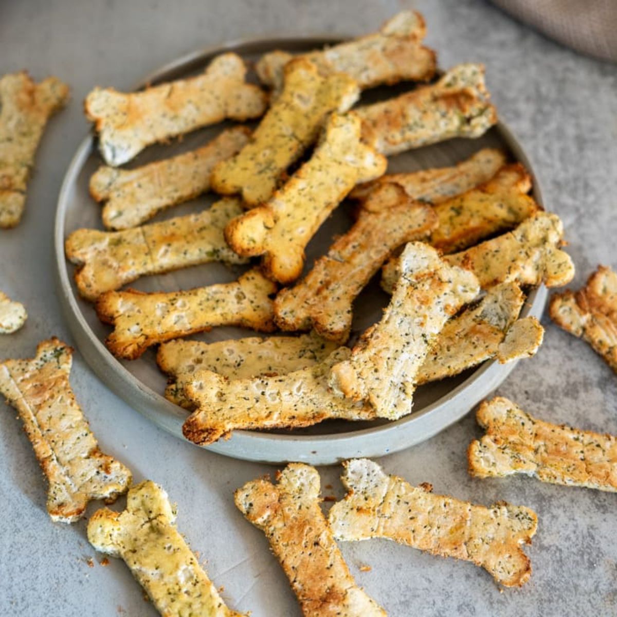 A plate is filled with bone-shaped biscuits, some scattered on a gray surface. The biscuits appear to be flavored with herbs.