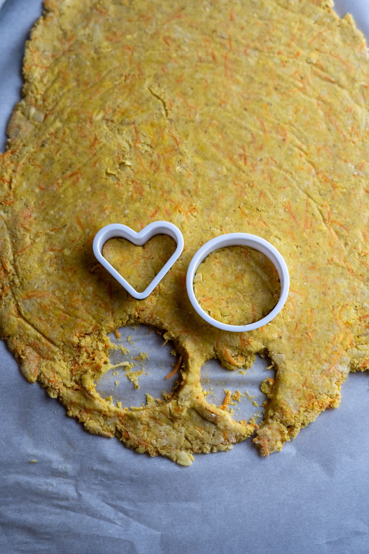 Dough rolled out on parchment paper with heart and circle cookie cutters, ready to create delightful Easter cookies for dogs, with one heart-shaped cut already made.