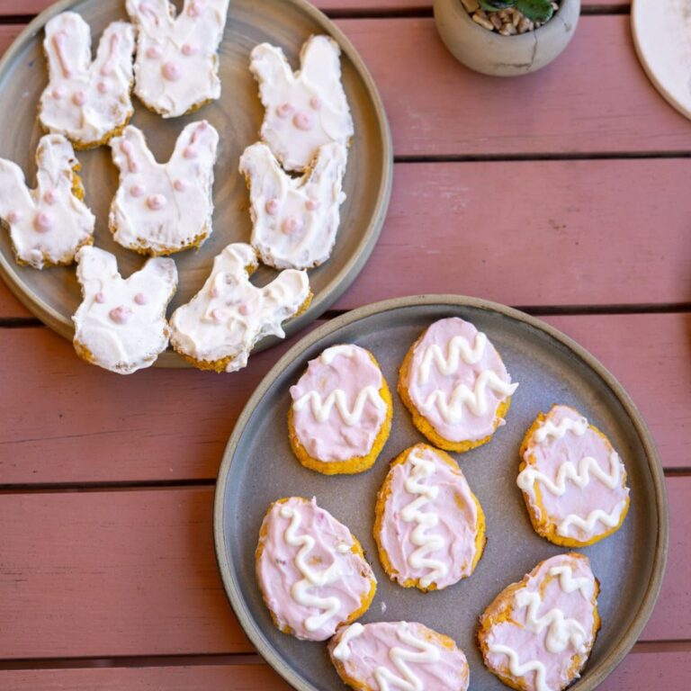Two plates of Easter-themed cookies on a pink wooden table. Top plate with bunny-shaped cookies with white icing, bottom plate with egg-shaped cookies with pink icing and white zigzags.