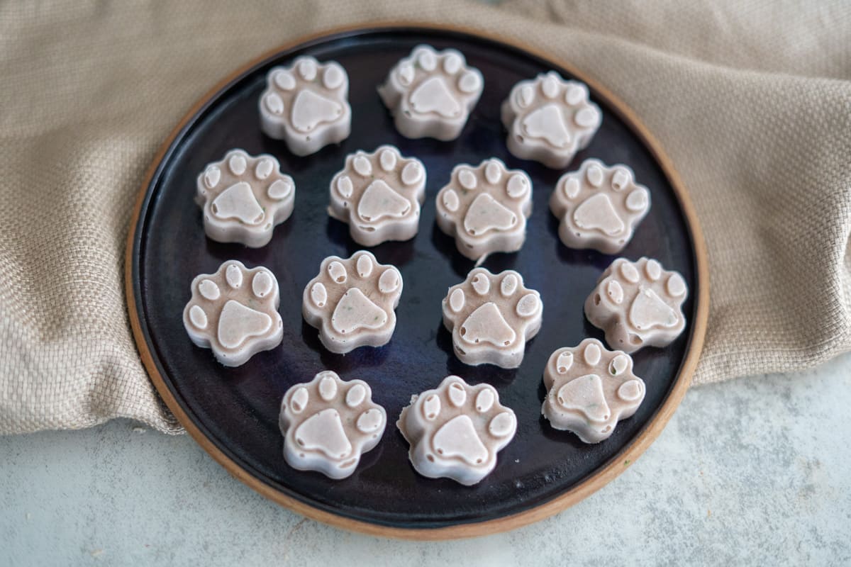 A round black plate holding fifteen paw-shaped, light brown treats with white icing, arranged in neat rows, set on a beige cloth.