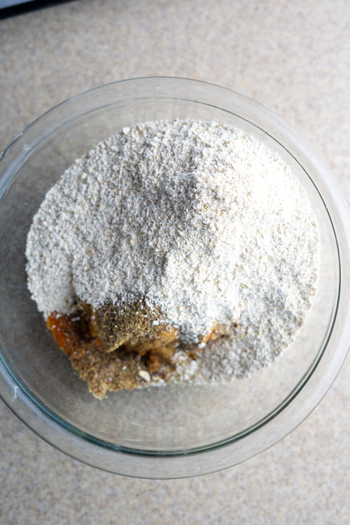 A glass bowl containing a dry flour and spice mixture for no-bake pumpkin dog treats sits on a light countertop.