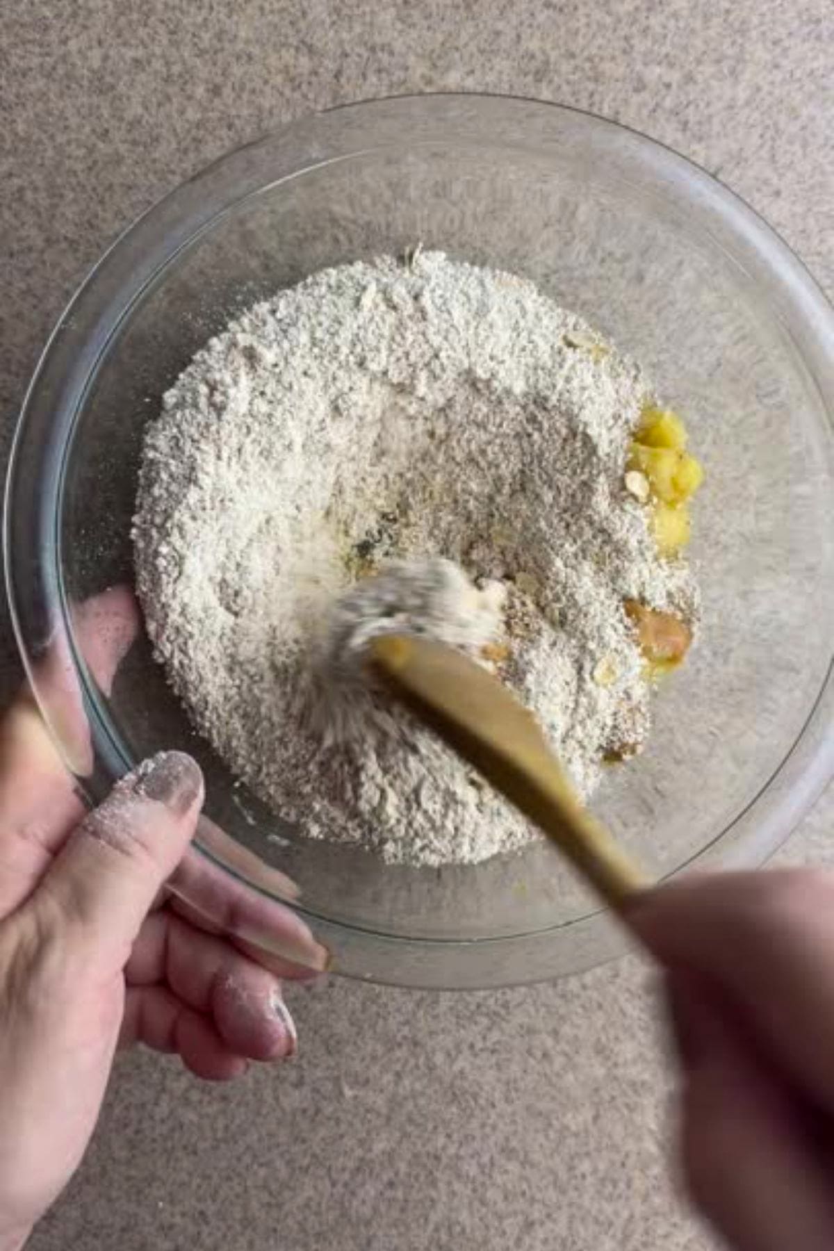 Person mixing flour and ingredients in a clear glass bowl using a wooden spoon on a countertop.