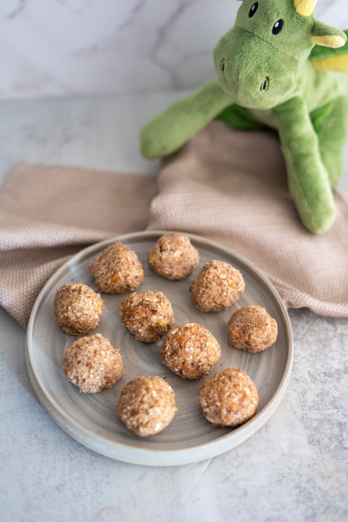A plate of bite-sized, no-bake banana dog treats rests on a light surface, with a green stuffed dragon toy playfully perched in the background.