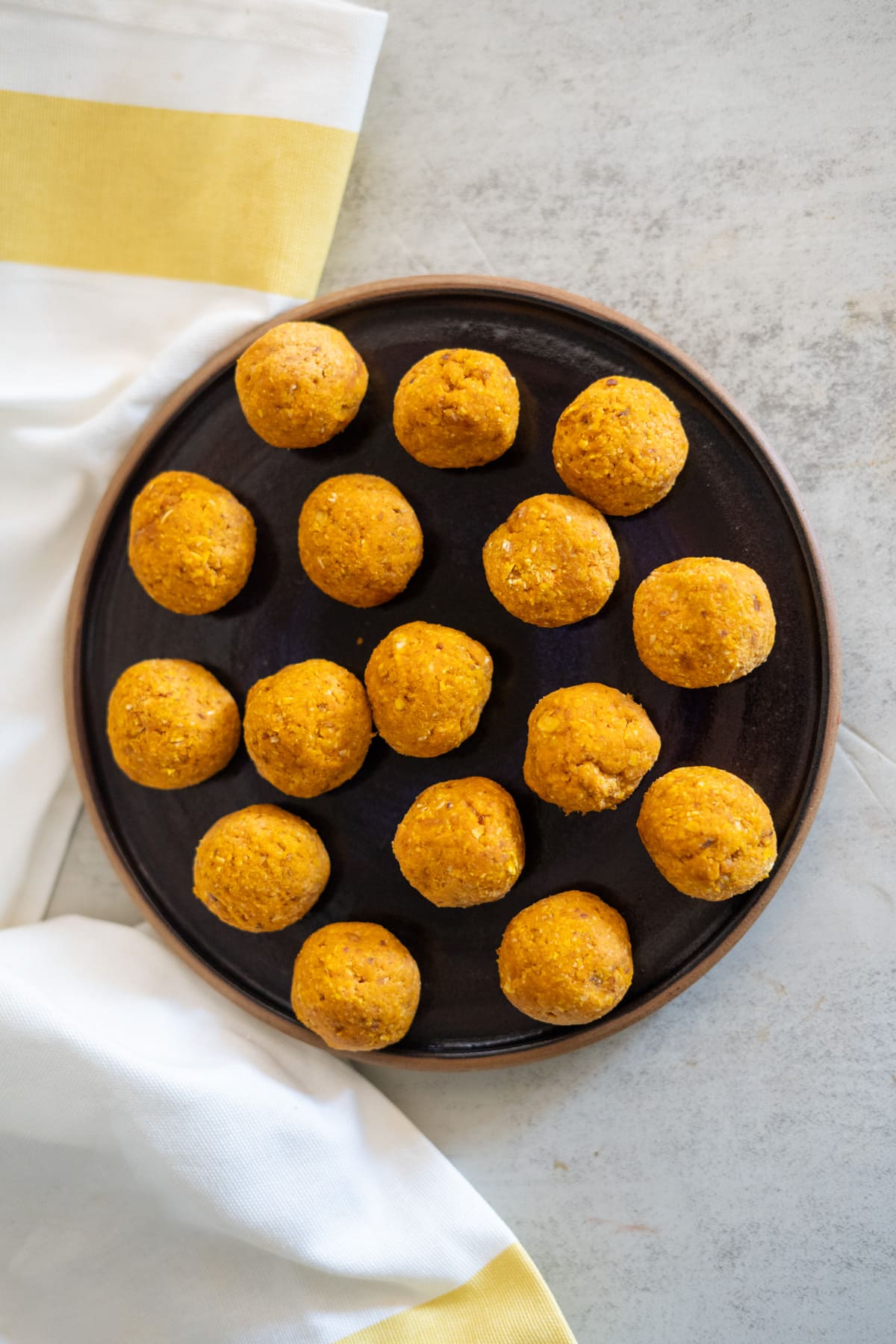 Round orange balls resembling no-bake pumpkin dog treats are arranged on a dark plate, placed on a light surface with a white and yellow cloth partially visible.