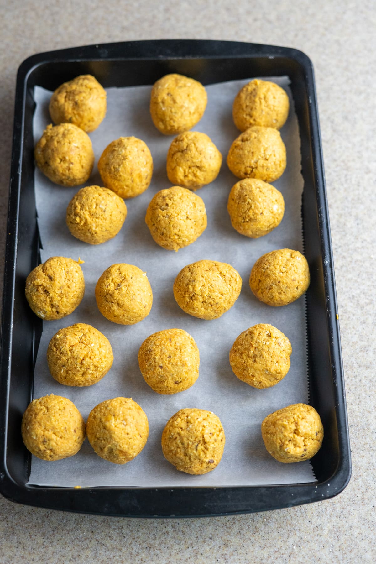 A baking tray with parchment paper holds 20 evenly spaced, round yellow dough balls, perfect for no-bake pumpkin dog treats, placed on a speckled countertop.