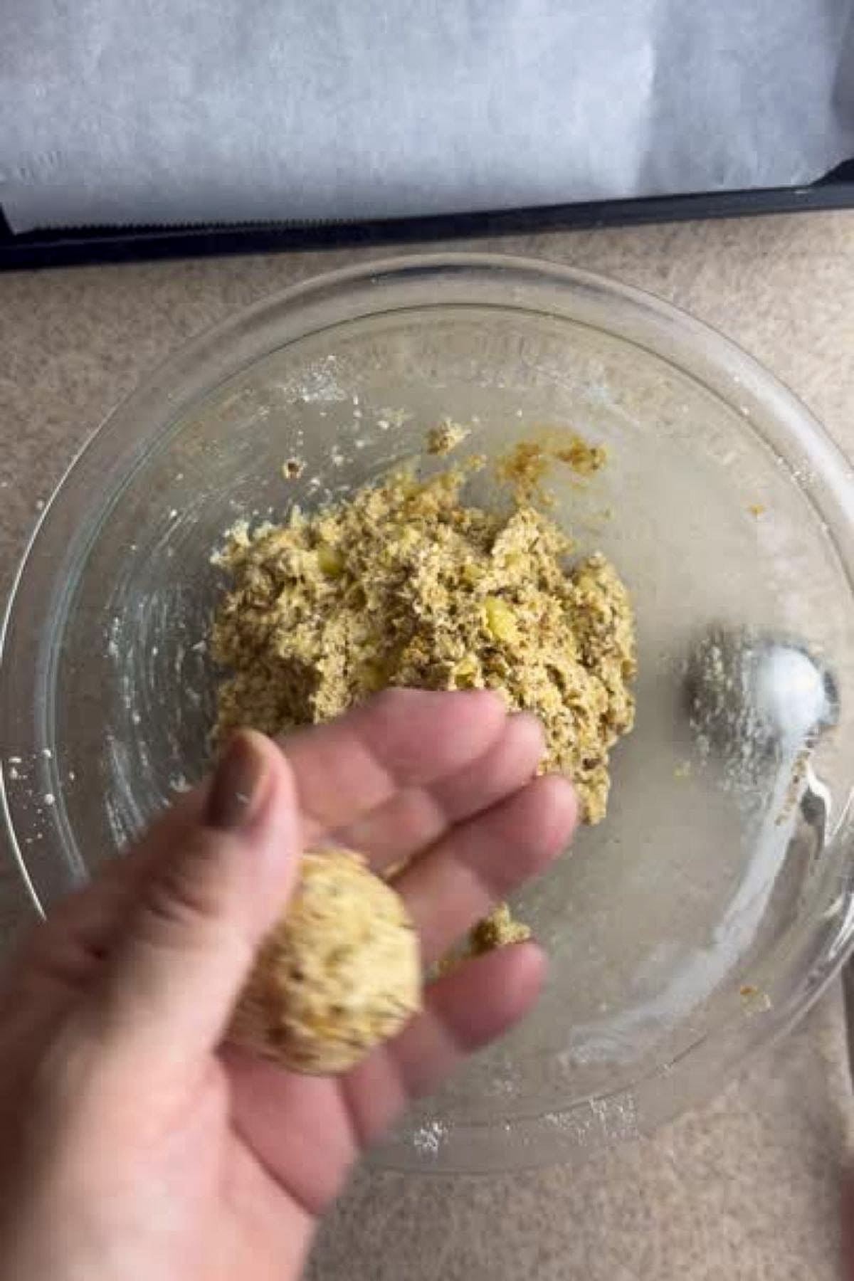 Hand shaping a ball of dough above a glass mixing bowl containing more dough on a kitchen counter.