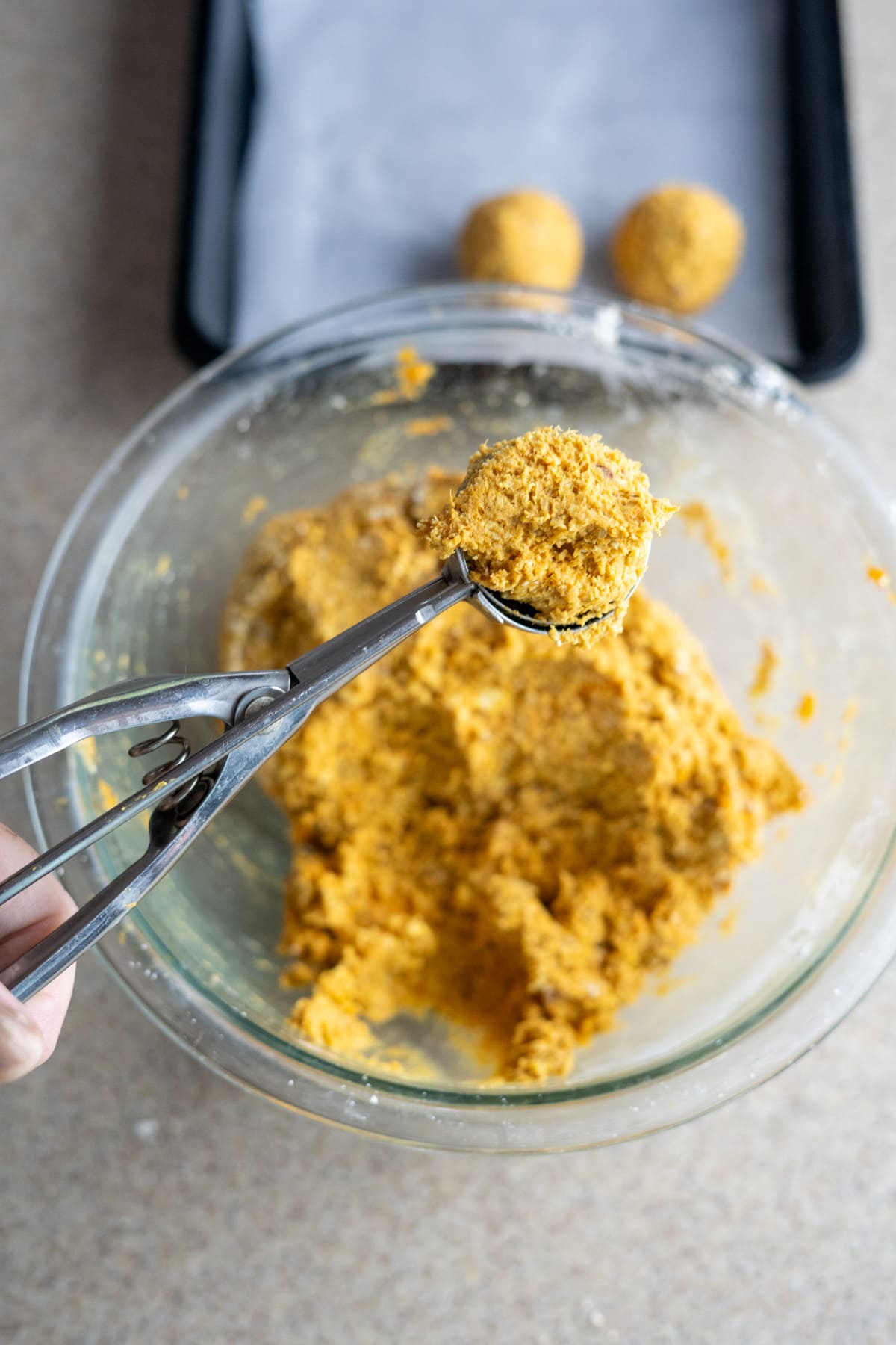 A person uses an ice cream scoop to portion dough for no bake pumpkin dog treats from a glass mixing bowl. Two dough balls rest on a baking sheet in the background.