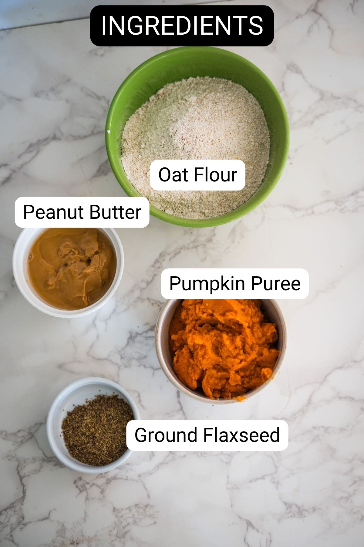 Labeled ingredients on a marble surface: oat flour in a green bowl, peanut butter in a white cup, pumpkin puree in a bowl, and ground flaxseed in a small dish.