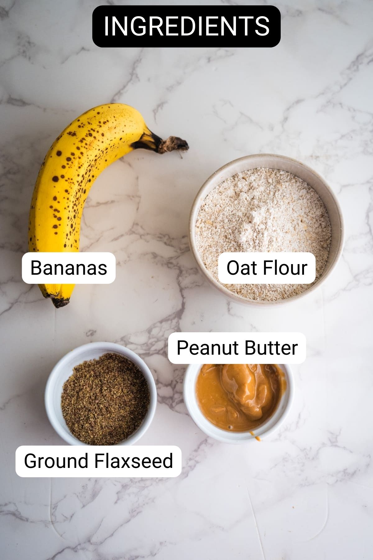 Ingredients on a marble surface: a ripe banana, a bowl of oat flour, a bowl of peanut butter, and a bowl of ground flaxseed.
