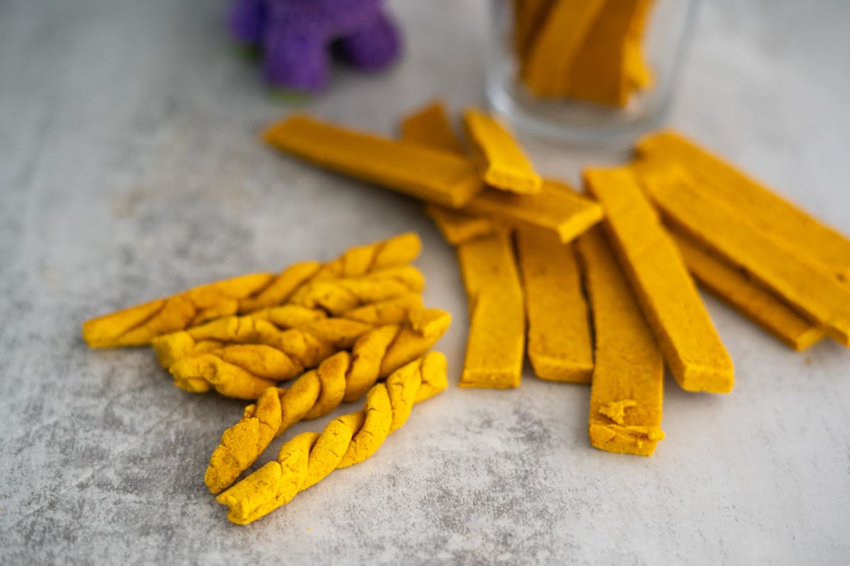 A close-up of yellow dog treats, some twisted and some rectangular, on a gray surface with a glass jar in the background.
