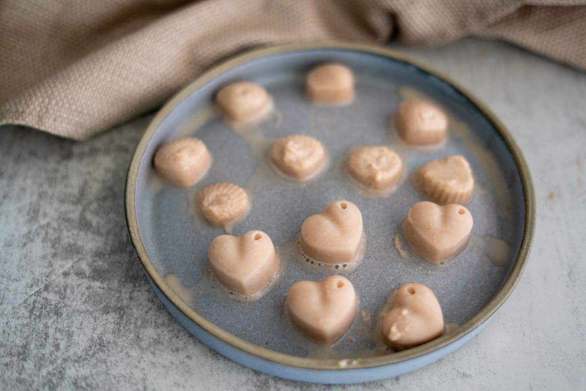A round plate holds several light brown, heart-shaped and shell-shaped candies or treats on a grey surface with a beige cloth in the background.