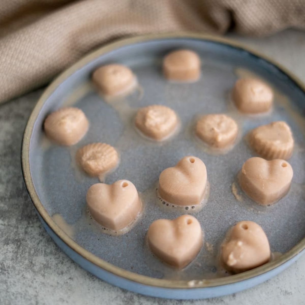 A plate with heart-shaped and round frozen treats in light brown color, partially melted, on a gray surface with a beige textured cloth in the background.
