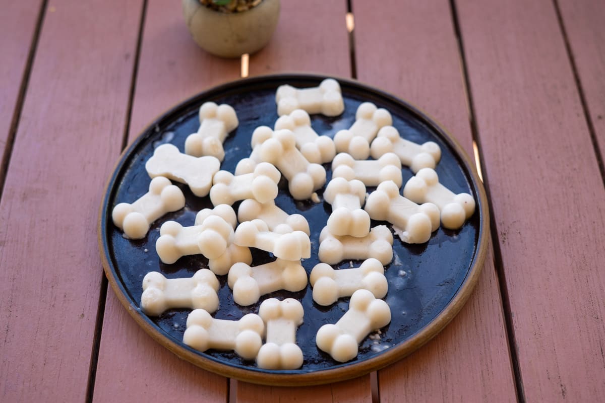A round black plate on a wooden table holds many small, white, bone-shaped treats. A small potted plant is in the background.