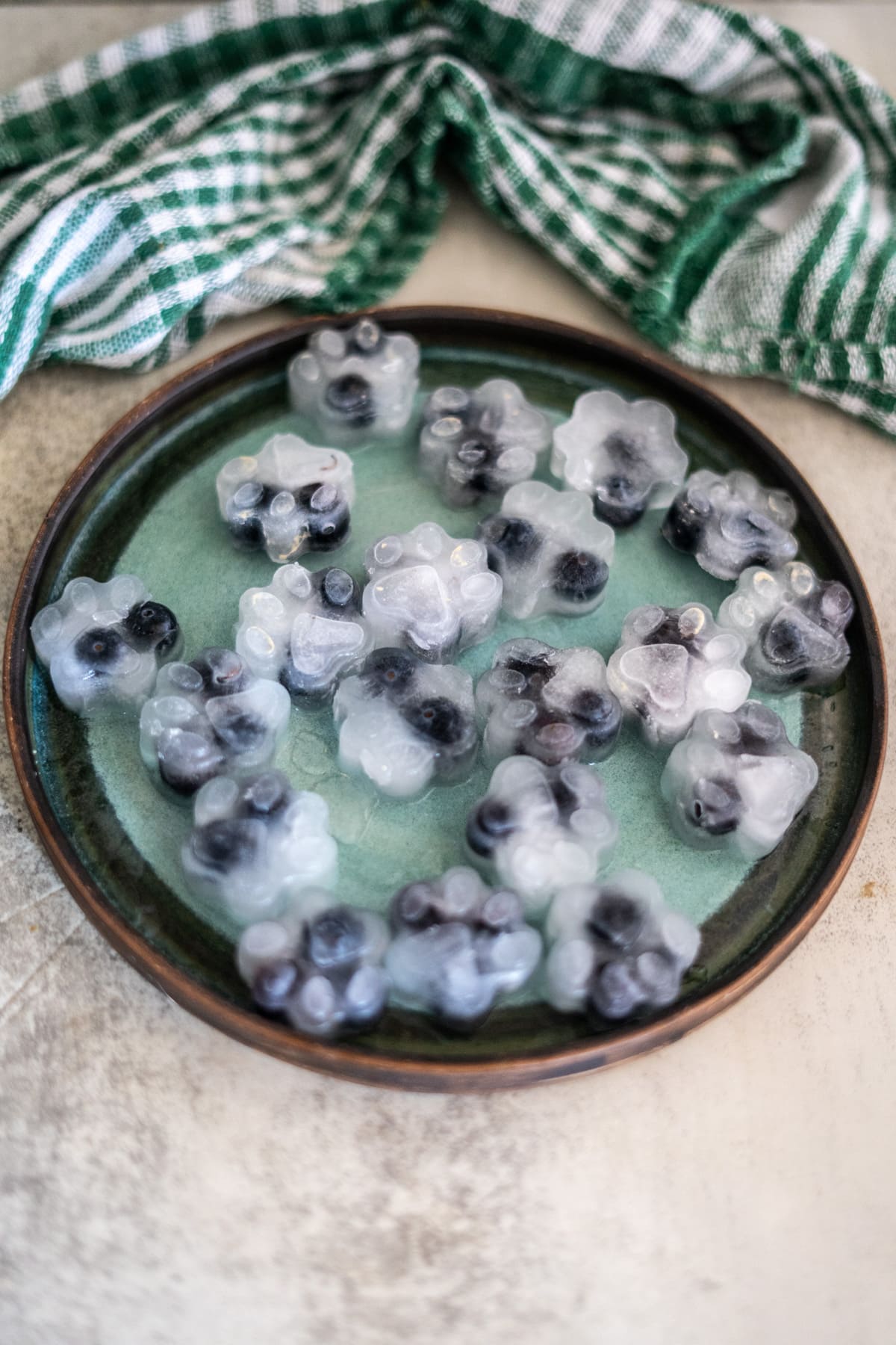 A green plate with paw-shaped coconut water blueberry dog treats, crafted as ice cubes with blueberries inside, sits on a light surface with a green and white checked cloth in the background.