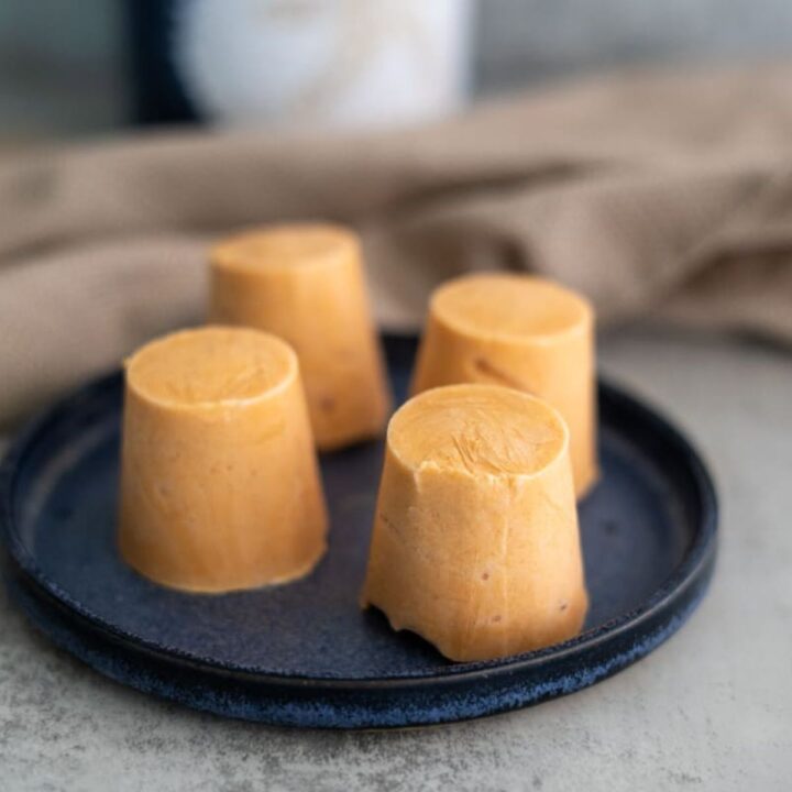 Four cylindrical pieces of orange-colored frozen dessert are arranged on a dark blue plate, with a brown cloth and a blurred background.