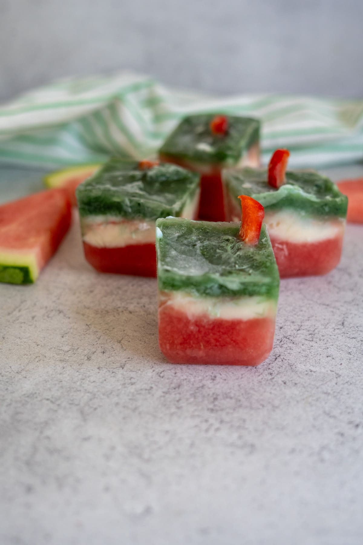 Three-layered watermelon frozen dog treats resembling watermelon slices with red, white, and green layers, garnished with small red pepper pieces on a light grey surface.