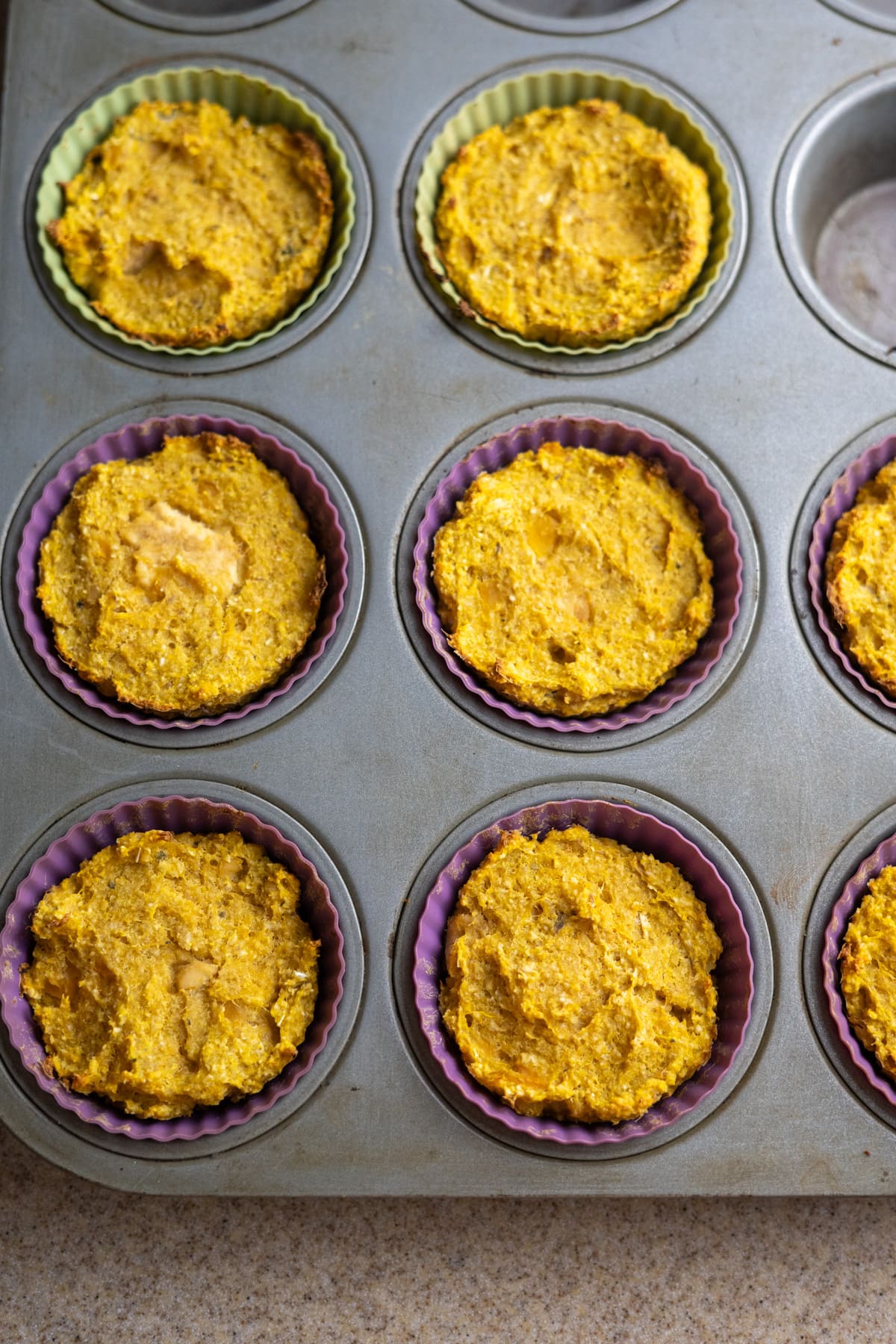 A muffin tin with seven baked sweet potato pupcakes in silicone liners, with one empty compartment, placed on a kitchen countertop.