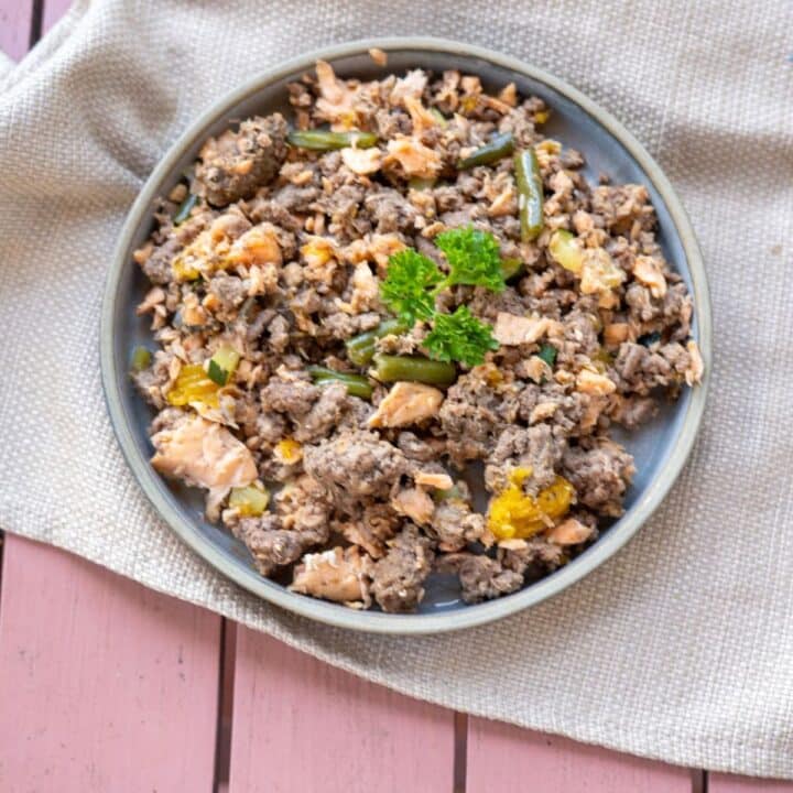 A bowl of mixed ground meat, green beans, and yellow bell peppers, garnished with parsley, placed on a gray napkin over a pink wooden surface.