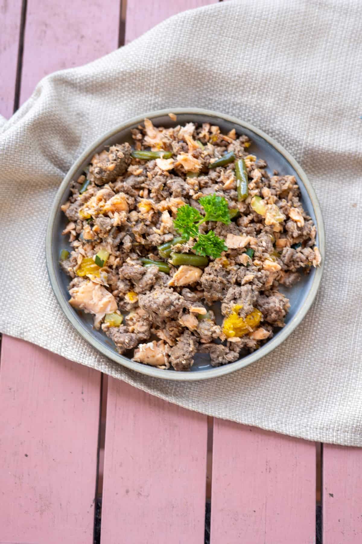 A plate of mixed cooked ground meat, vegetables, and rice, garnished with parsley, on a beige cloth atop a pink wooden surfaceโperfectly suited as a wholesome salmon meal for dogs.
