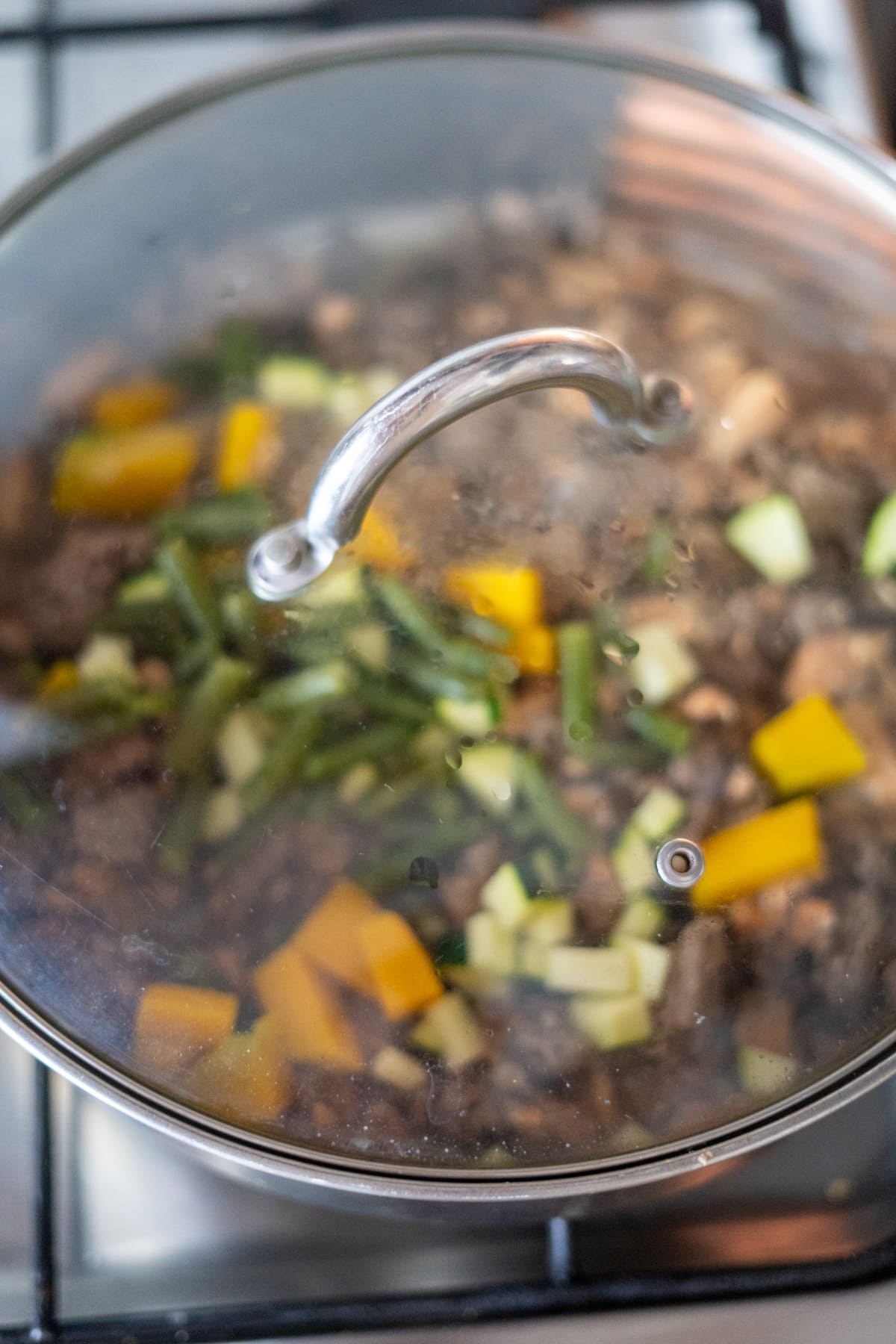 A glass-lidded pot on a stove contains chopped vegetables and food steaming inside.