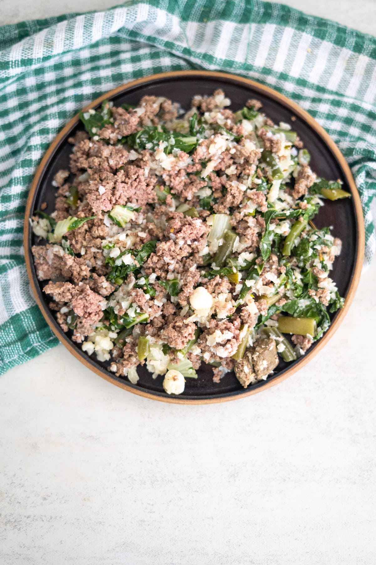 A plate of ground beef mixed with green vegetables and cauliflower rice for dogs is placed on a light surface next to a green and white checkered cloth.