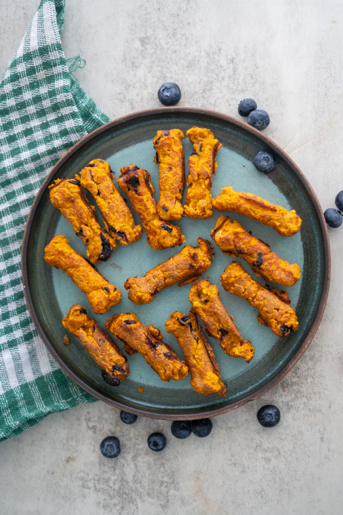 A plate with twelve bone-shaped Dash Dog Treats, arranged in a circle, sits on a light surface with scattered blueberries and a green checkered cloth nearby.