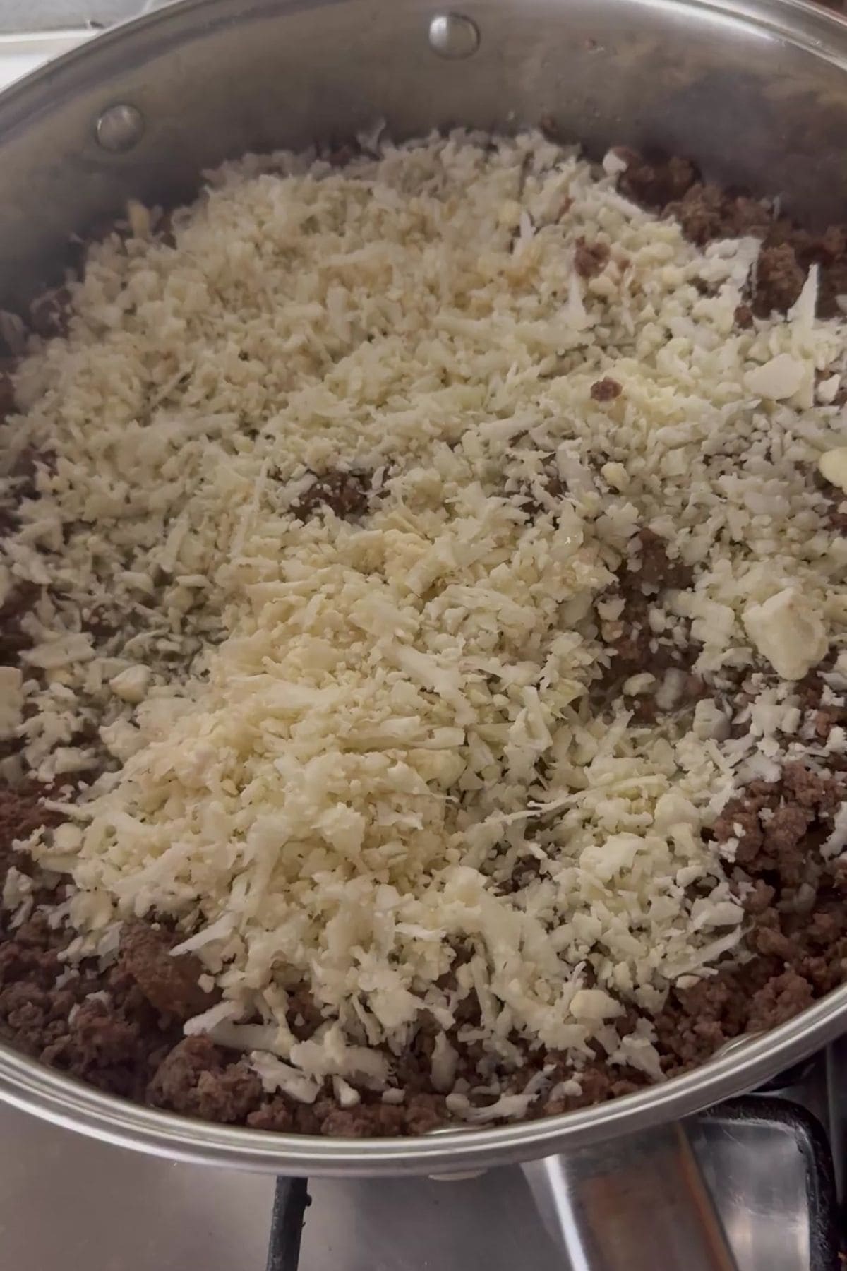 Ground beef in a skillet topped with a layer of shredded cheese, cooking on a stovetop.