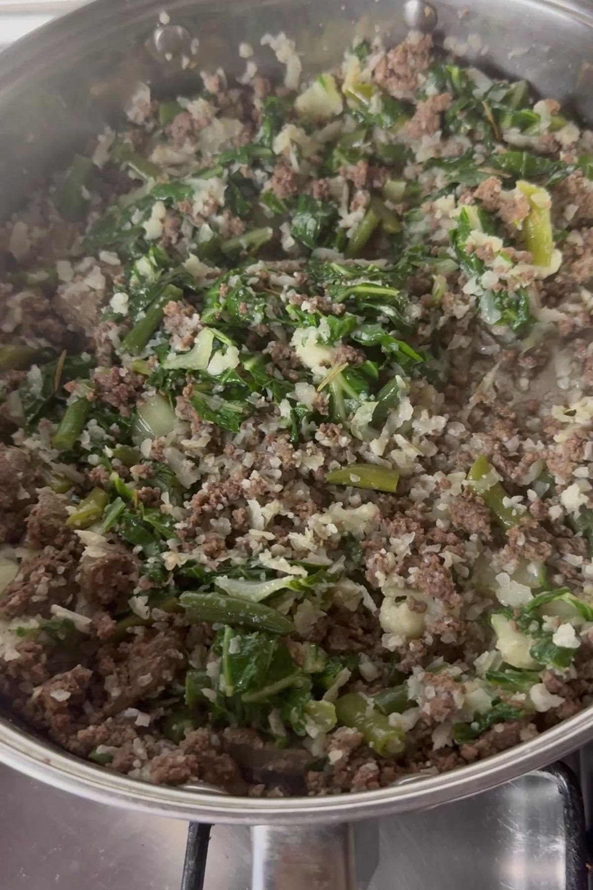 A stainless steel pan on a stove contains a mixture of ground beef, chopped greens, cauliflower rice, and diced onions being cooked.