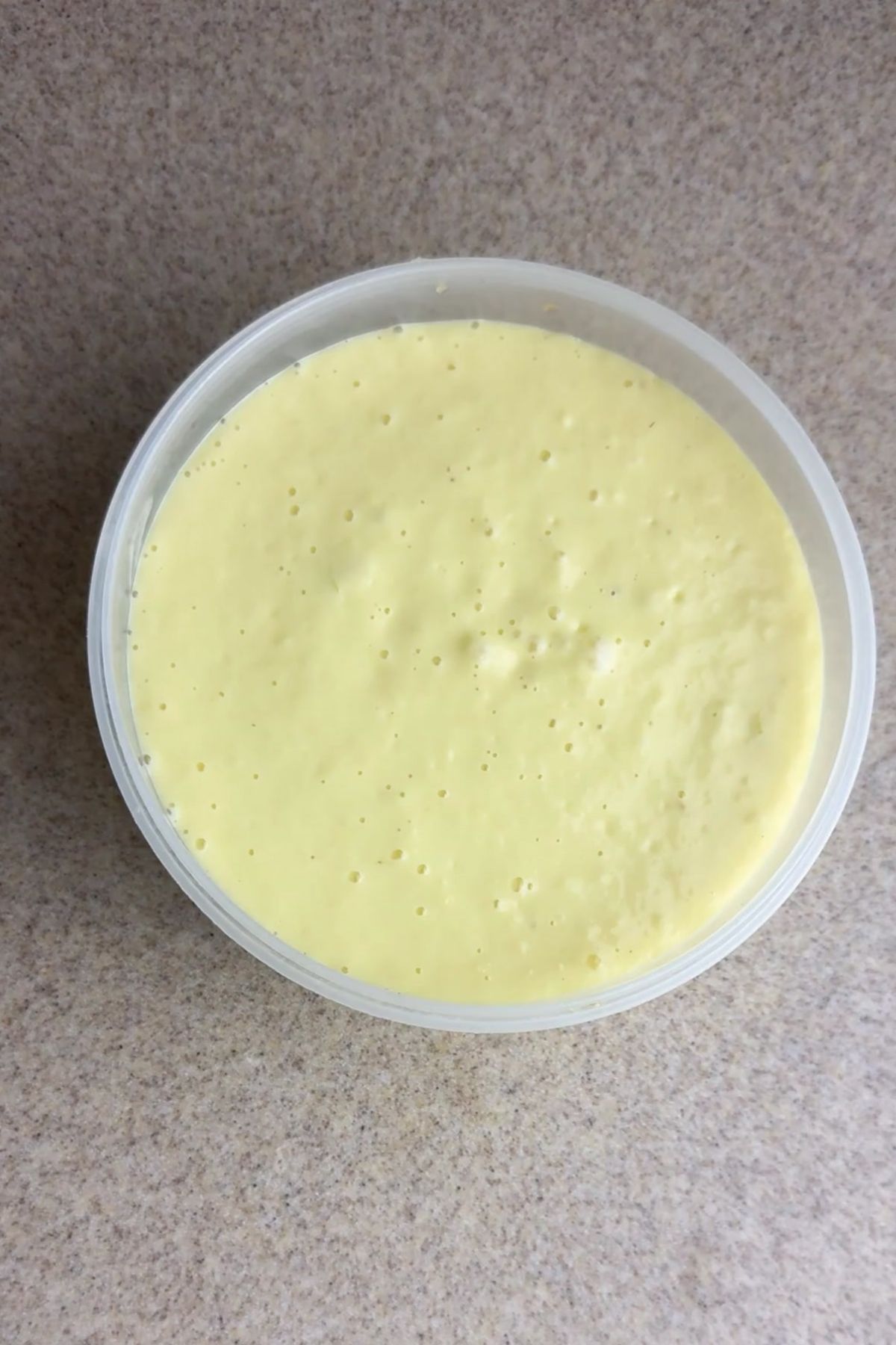A round plastic container filled with a smooth, pale yellow, creamy substance sits on a speckled countertop.
