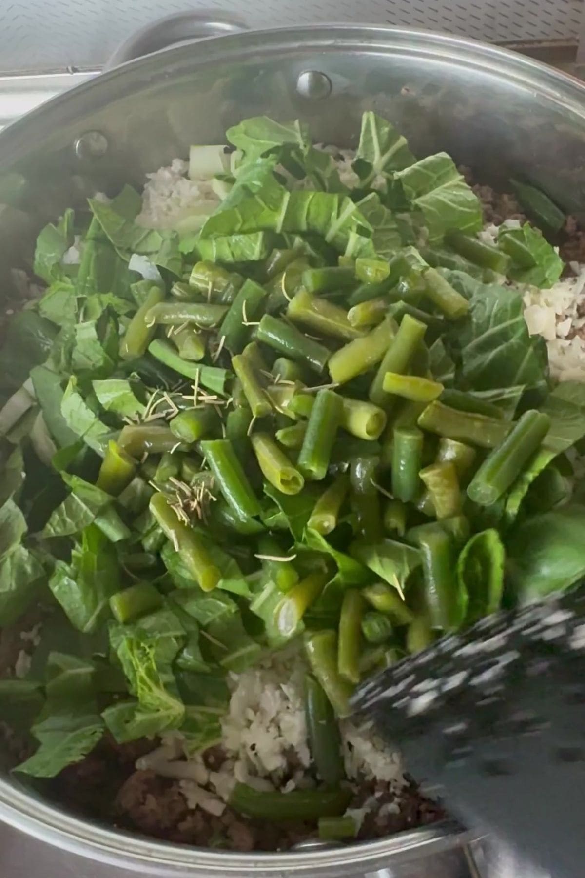 Chopped greens and green beans are being added to a pan of rice and ground meat on a stovetop.