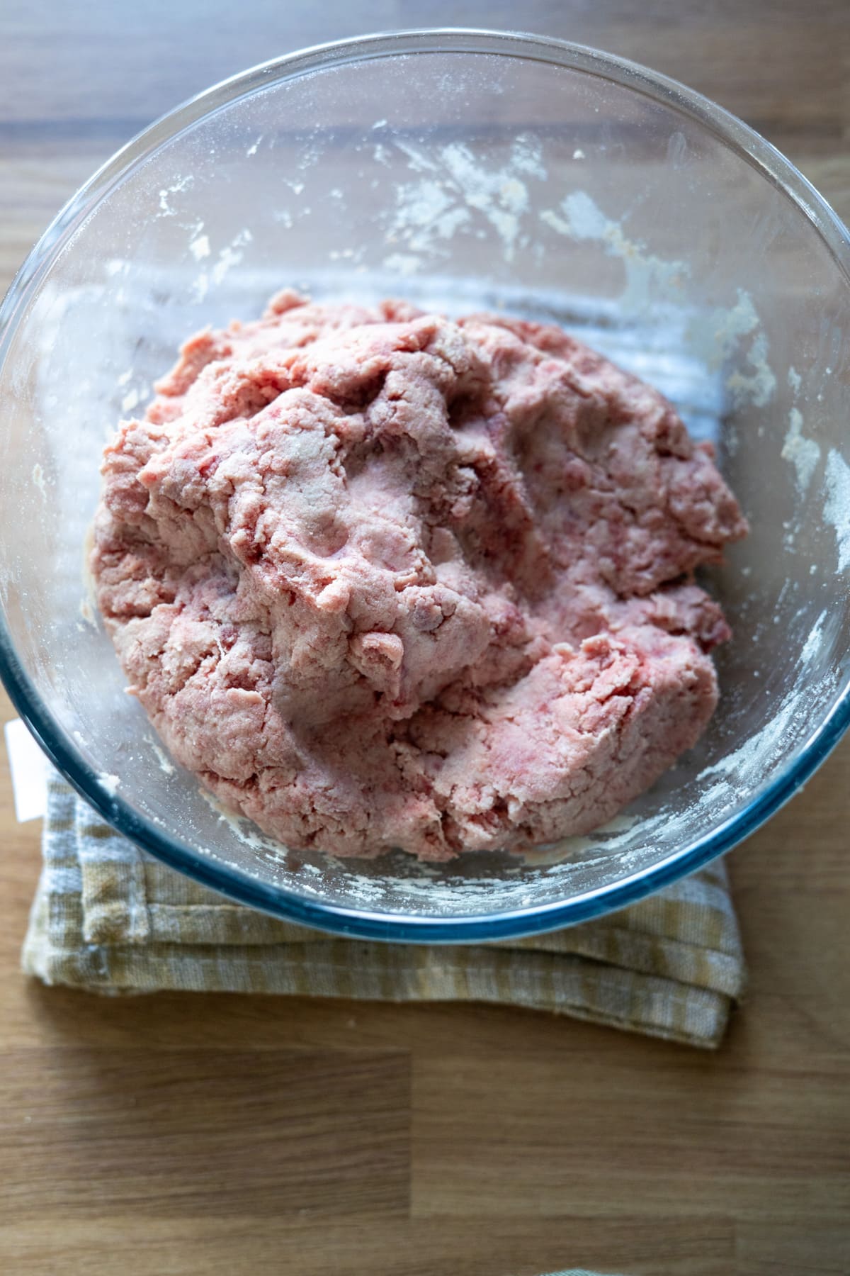 A glass bowl containing a lump of raw, pink dough for beef dog treats sits on top of a folded kitchen towel on a wooden surface.