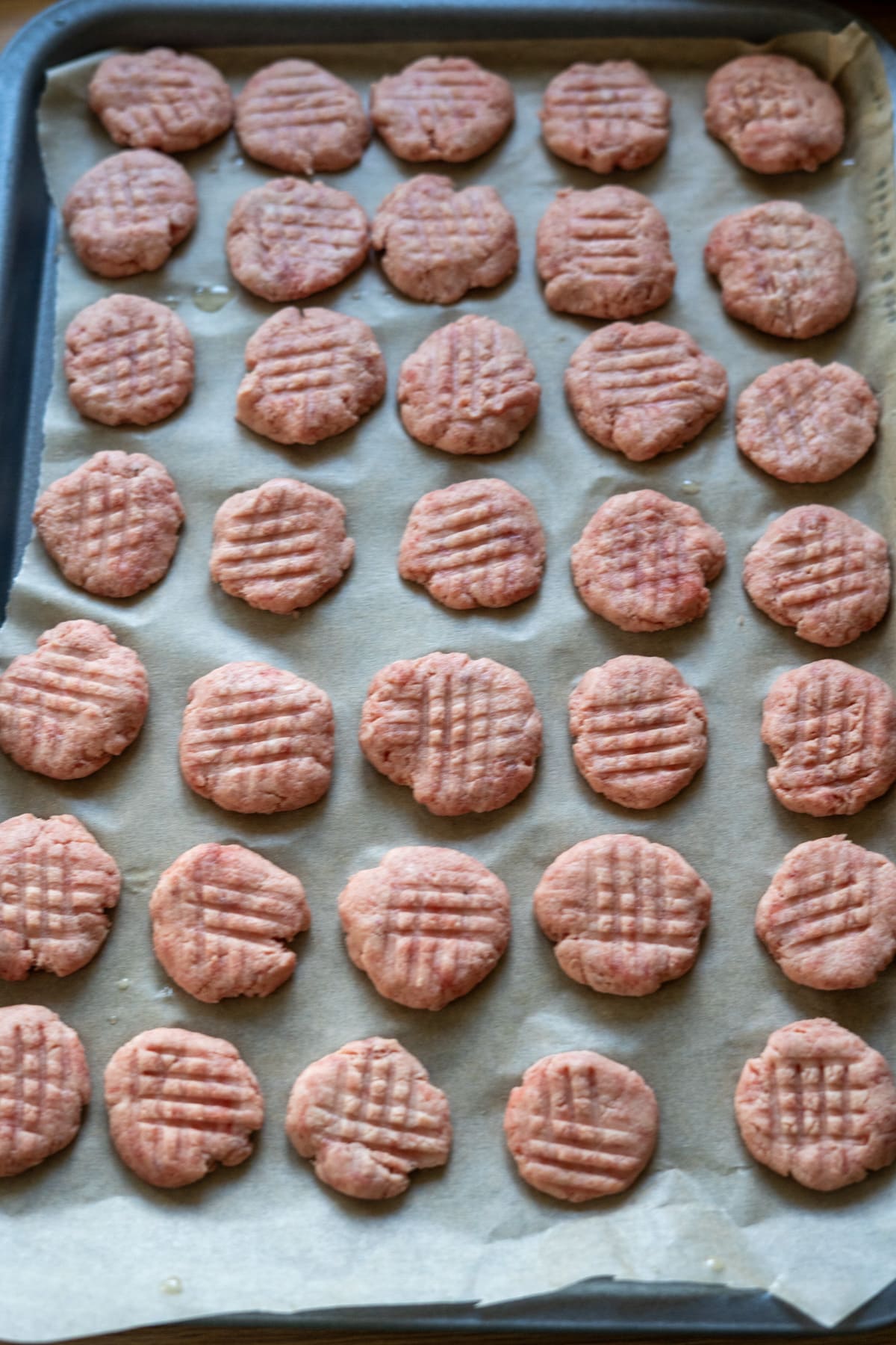 A baking sheet lined with parchment paper holds rows of raw, round patties of beef dog treats, each pressed with a crisscross pattern on top.