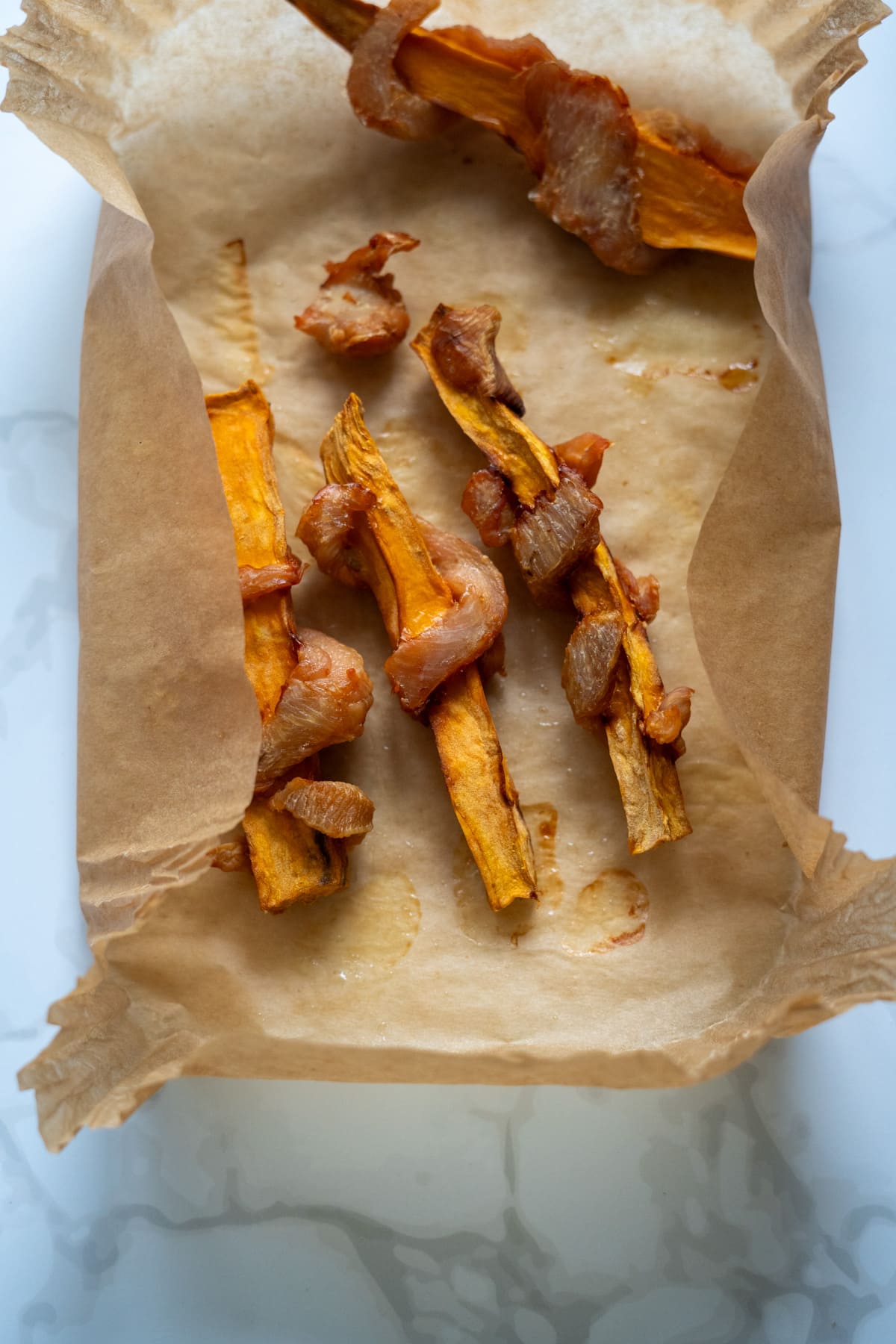 Baked sweet potato wedges wrapped with pieces of bacon on parchment paper in a baking dish.