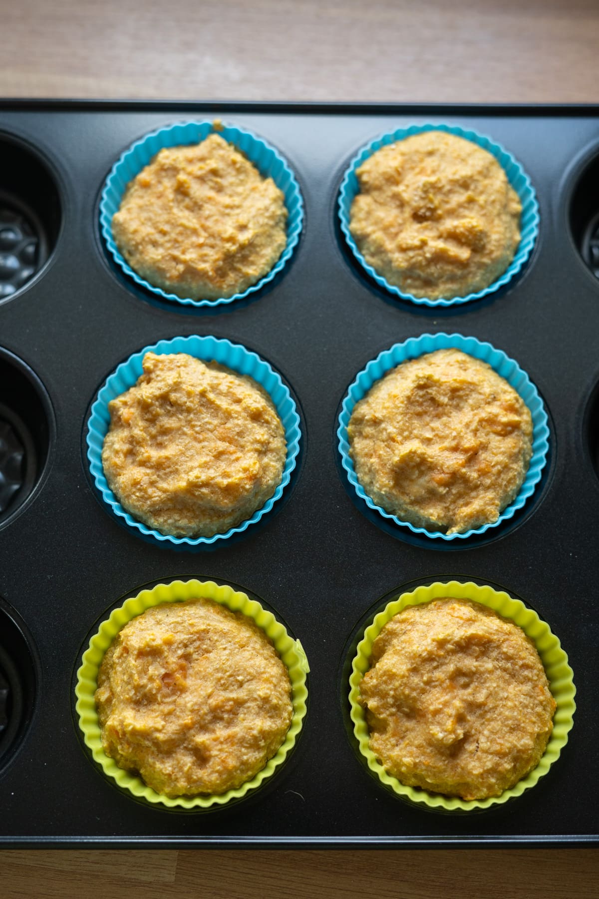 Six unbaked muffins in blue and green silicone cupcake liners arranged in a black muffin tray, sitting on a wooden surface.