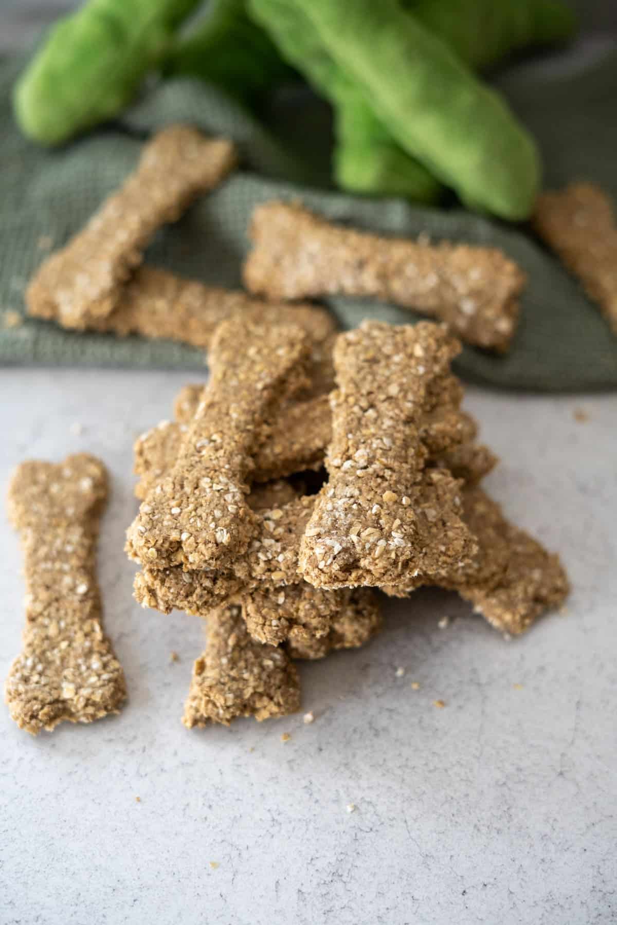 A pile of homemade dog biscuits shaped like bones sits on a light surface, with more biscuits and a green cloth in the background.