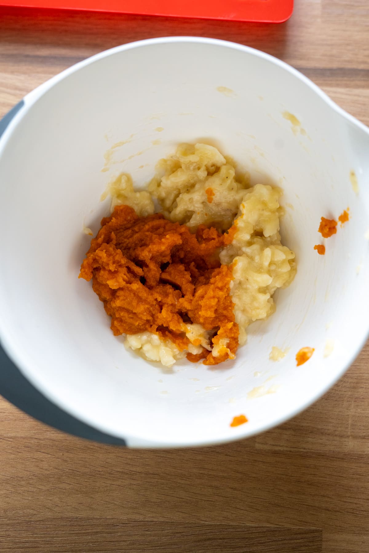 A white mixing bowl containing mashed bananas and pumpkin puree on a wooden surface.