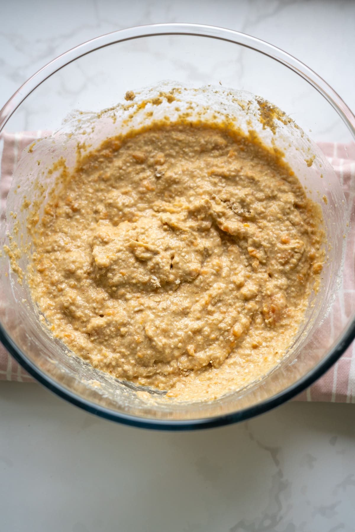 A glass bowl filled with a thick, lumpy batter sits on a pink-and-white striped cloth on a marble countertop.