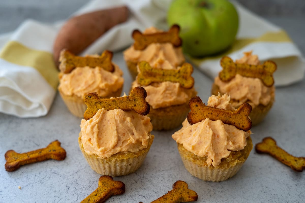 Five cupcakes with orange frosting, each topped with a small bone-shaped biscuit, are arranged on a light surface with an apple and kitchen towel in the background.