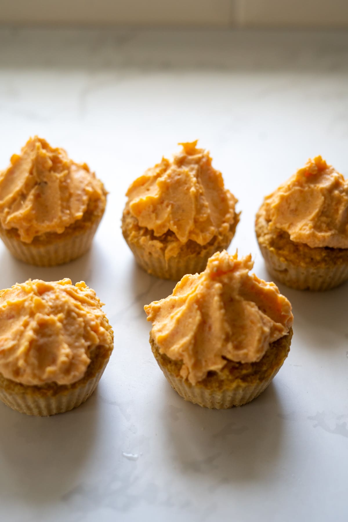 Five cupcakes with orange frosting arranged on a white marble surface.