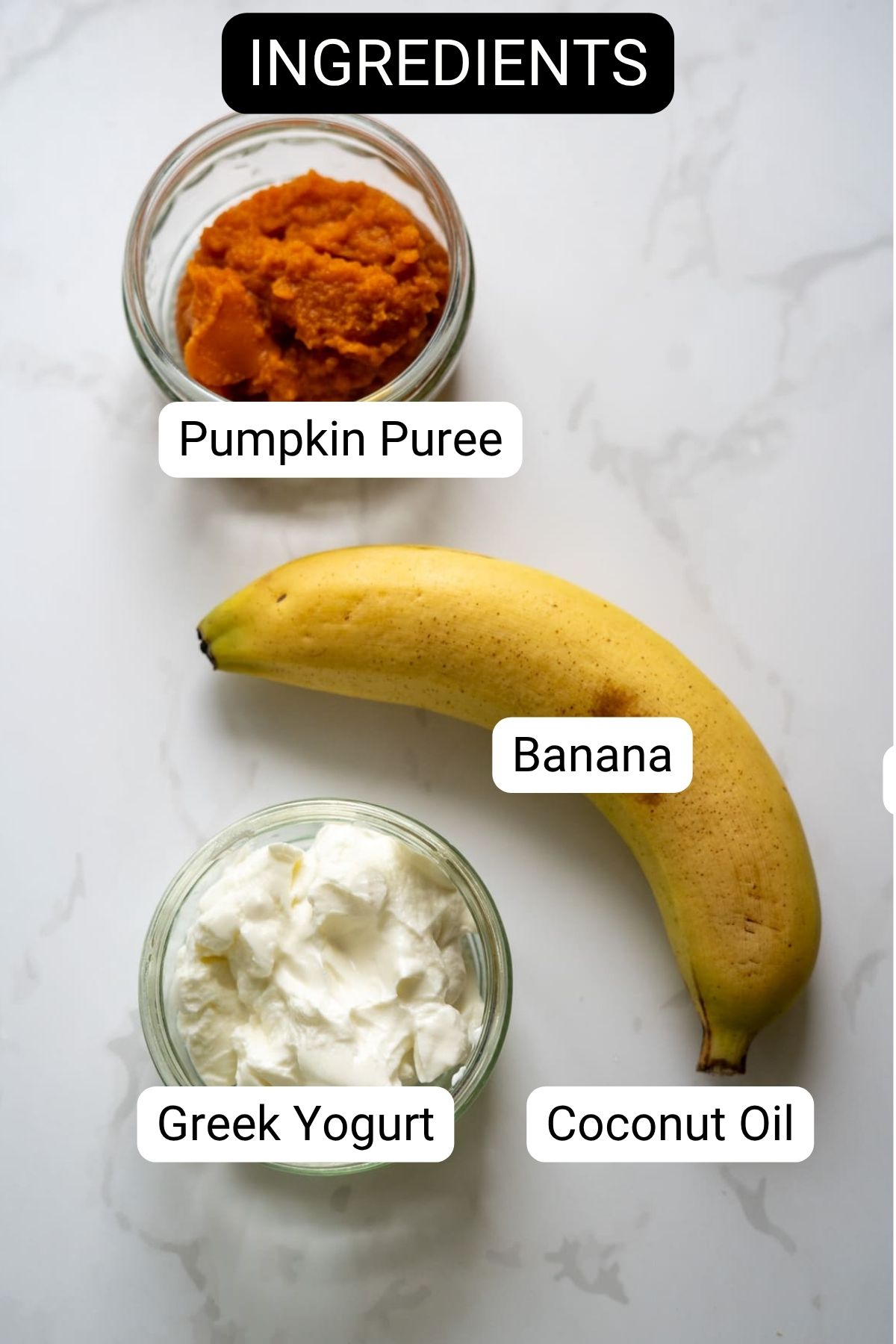 A banana, a bowl of pumpkin puree, a bowl of Greek yogurt, and a bowl of coconut oil are arranged on a white surface, each labeled with text.