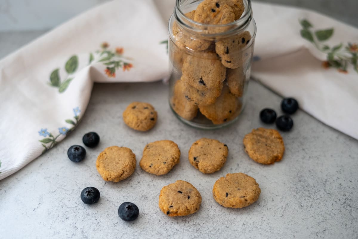 A glass jar of small round cookies sits on a stone surface, with several cookies and scattered blueberries around it. A white floral cloth is in the background.