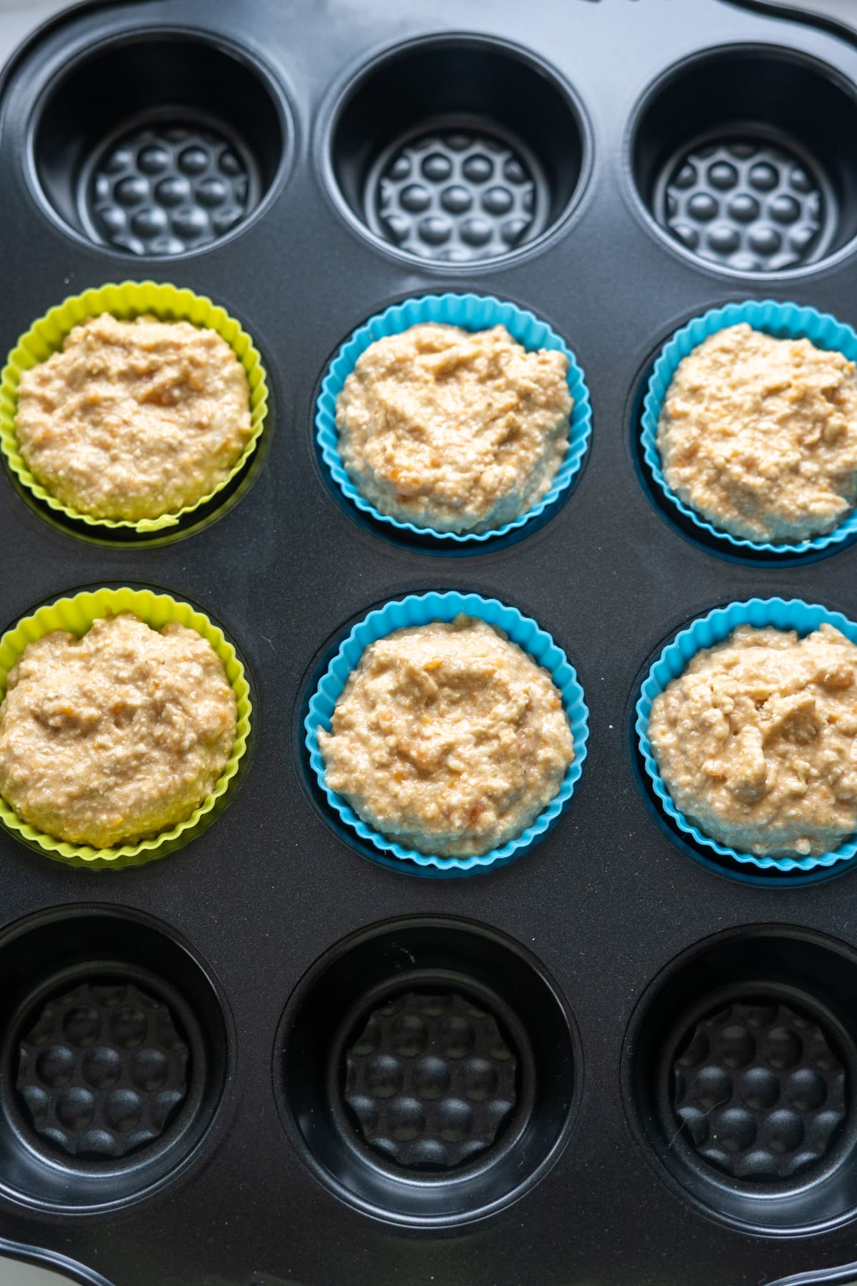 A muffin tin with six silicone liners, each filled with muffin batter, and six empty spaces visible.