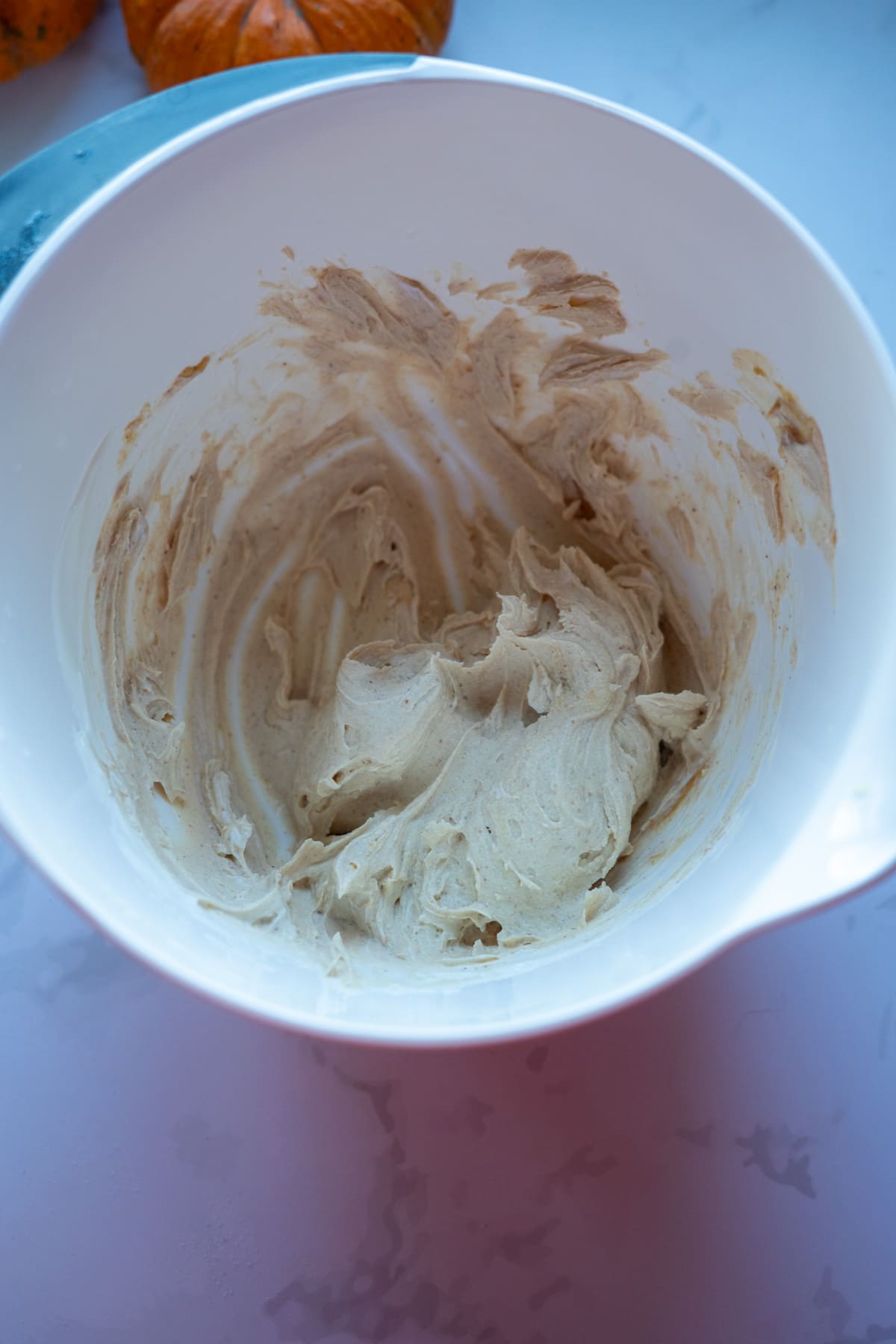 A white mixing bowl containing light brown whipped batter for pumpkin pupcakes sits on a marble countertop.
