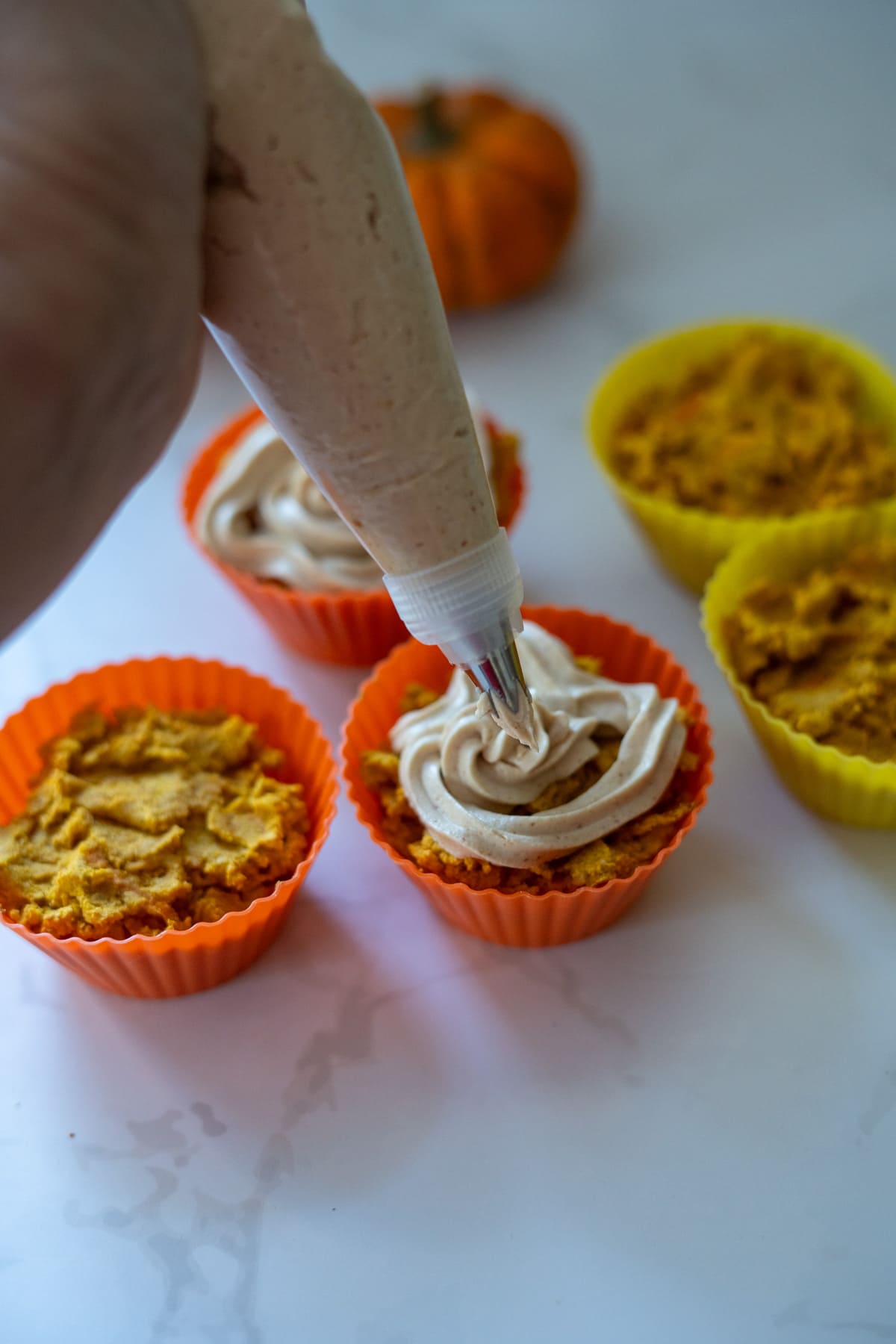A hand uses a piping bag to add frosting to pumpkin pupcakes in orange and yellow silicone baking cups on a white surface.